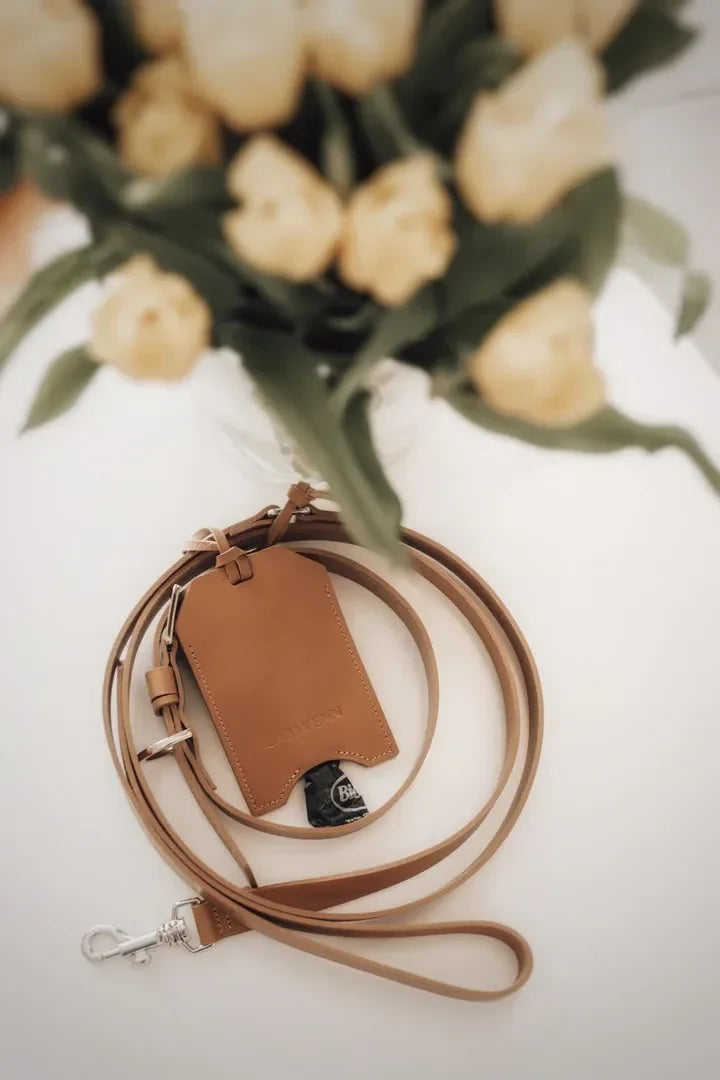 Distributeur de sacs pour chien en cuir naturel, posé près de fleurs sur une table claire, style artisanal.