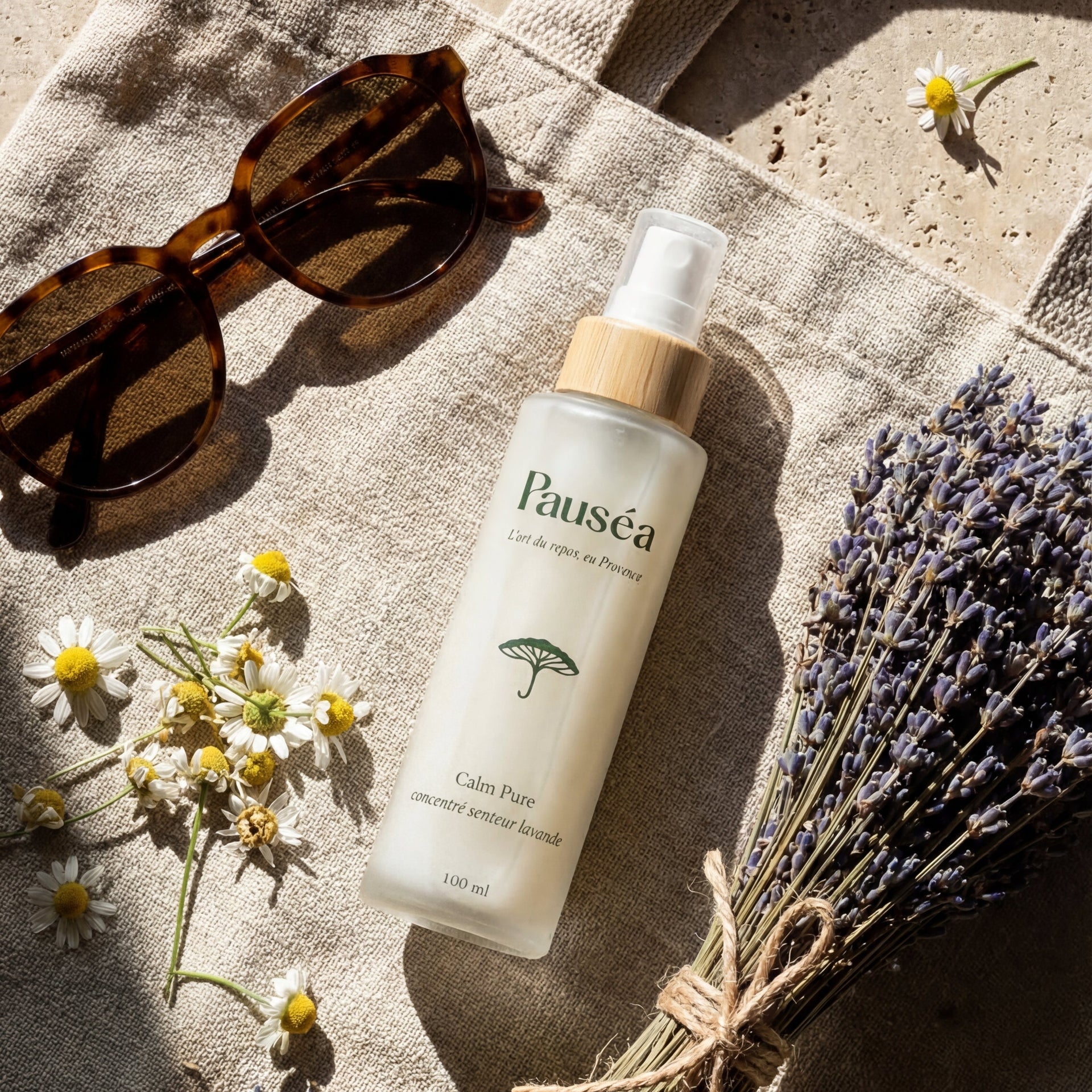 Composition authentique avec le concentré de lavande Pauséa fait main, un bouquet de lavande séchée de Provence, des fleurs de camomille et des lunettes de soleil sur un sac en lin naturel.